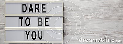 `Dare to be you` on a lightbox, cliboard with blank sheet of paper on a white wooden background, top view. Flat lay, overhead, Stock Photo