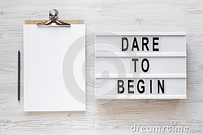 `Dare to begin` words on a lightbox, clipboard with blank sheet of paper on a white wooden background, top view. Overhead, from Stock Photo
