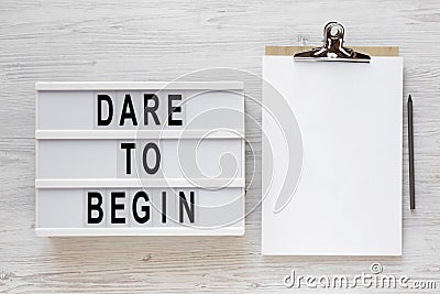 `Dare to begin` words on a lightbox, clipboard with blank sheet of paper on a white wooden surface, top view. Overhead, from abo Stock Photo