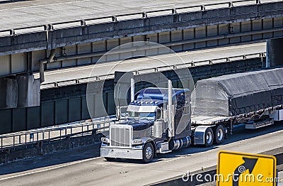 Dark blue classic big rig semi truck transporting covered cargo on flat bed semi trailer running on multilevel overpass highway Stock Photo