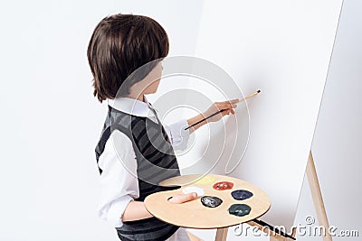 The dark-haired boy draws on the easel. He holds the paint and brush in his hand. Stock Photo