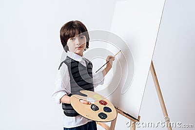 The dark-haired boy draws on the easel. He holds the paint and brush in his hand. Stock Photo