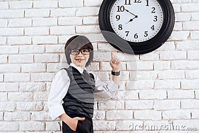 The dark-haired boy is standing by the white wall in a suit. He points at the wall clock. A small future businessman. Stock Photo