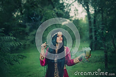 Dark-haired enchantress of ancient tribe creates a new magic spell, holds a golden bowl with mist and haze in her hands Stock Photo