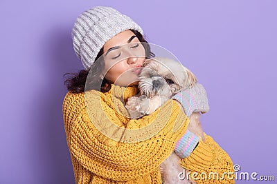 Dark haired girl wearing warm yellow sweater and cap kissing her little dog, blithesome european woman expressing love to her Stock Photo