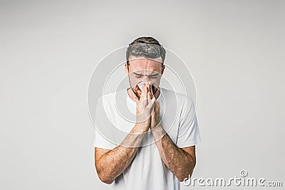 Dark-haired man is standing near the white wall and sneezing. Seems like he caught some cold and soon will be very ill Stock Photo