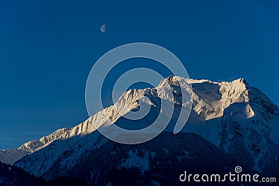 Dark mountain with sunlit snowy top - the moon is visable in early afternoon Stock Photo