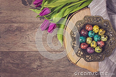 Dark photo of Easter quail eggs in an ancient metal bowl. Beautiful spring flowers - purple tulips on a wooden background. Floral Stock Photo