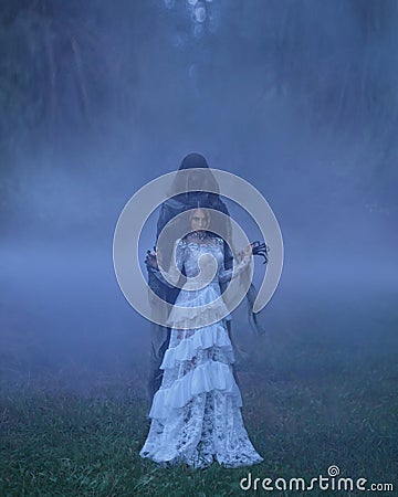 Dark Queen with neat hairdo in a white vintage dress and a silver necklace, standing in a forest full of thick purple Stock Photo