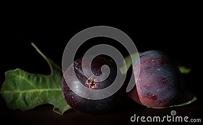 Dark shot of two red figs lying side by side on a table. A few fig leaves in the background. The room is dark Stock Photo
