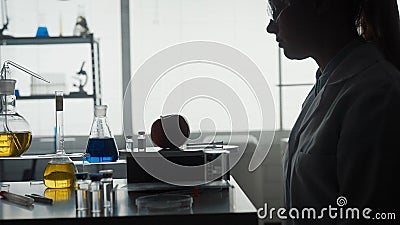 Dark silhouette of a female scientist sitting at a table in front of an apple, a syringe, and an ampoule of a chemical Stock Photo