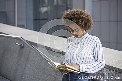 The dark-skinned girl is standing on the steps leaning on the railing. She is reading a book in the street near the office Stock Photo