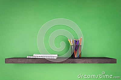 A dark wooden shelf against the background of a green wall on which is set a glass with pencils and notebooks with binding Stock Photo