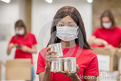 Darkskinned girl in protective mask with canned food Stock Photo