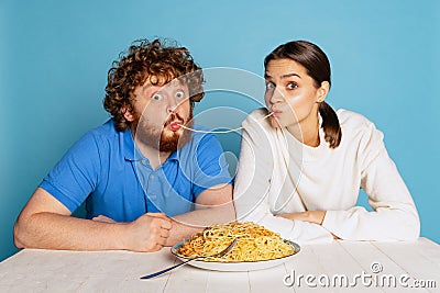 Cute young man and girl sitting together and tasting noodles, pasta isolated on blue studio background. Stock Photo