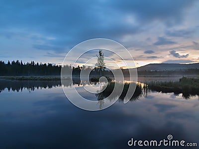 Daybreak autumn lake with mirror water level in mysterious forest, young tree on island in middle. Fresh green color of herbs Stock Photo