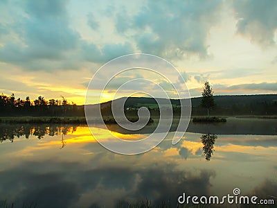 Daybreak autumn lake with mirror water level in mysterious forest, young tree on island in middle. Fresh green color of herbs and Stock Photo