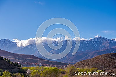 Daytime wide angle shot of Beautiful landscape of snow capped mountains and bushes and a village in the valley. Atlas, Morocco Stock Photo