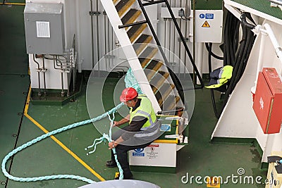 Deck hand fixes rope, whilst sat on deck of car ferry, with reflective jacket and red hat. Editorial Stock Photo