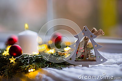 Decorative wooden Christmas tree with deer, burning white candle and fir wreath with red balls and warm garland lights near window Stock Photo