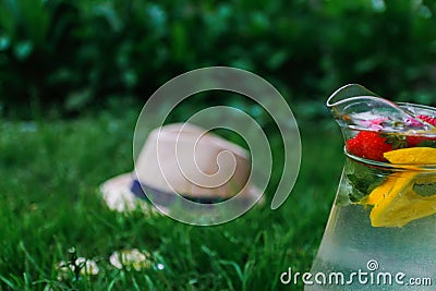 Defocus glass jug of lemonade with strawberry, slice lemon and leaves of mint in on blurred grass background. Pitcher of Stock Photo