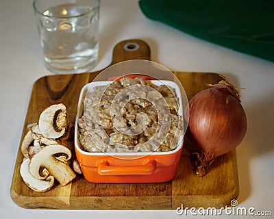 Delicious Beef stroganoff in a ceramic bowl decorated an onion, mushrooms a glass of water and a napkin on a cutting board Stock Photo