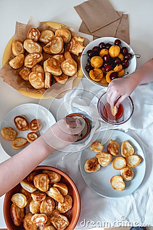 Delicious breakfast with tiny mini pancakes, cherries, apricots, jam on a white table. Small children hold pancakes in Stock Photo