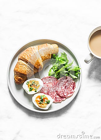 Delicious brunch - croissant, boiled egg, salami sausage, green salad and coffee with milk on a light background Stock Photo