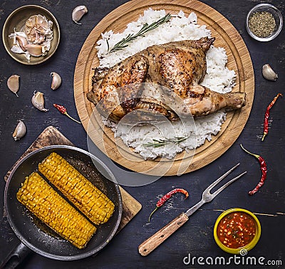 Delicious fried chicken with rice on a cutting board, fork for meat, spicy sauce, spices, garlic and corn in the pan on dark blue Stock Photo