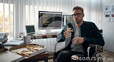 So delicious. Portrait of young hungry male web developer in a wheelchair eating pizza while sitting at his workplace in Stock Photo