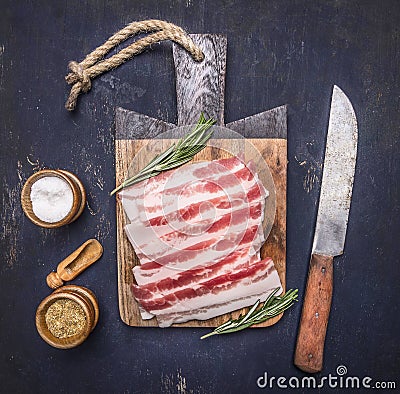 Delicious raw bacon, rosemary, knife and spices on a cutting board wooden rustic background top view close up Stock Photo