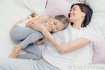 Delighted female child giggles joyfully as plays with her mother in comfortable bed, have positive smiles on faces, wear pyjamas, Stock Photo