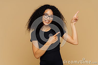 Delighted pleased dark skinned female points at upper right corner, shows place for your advertising content, wears glasses and Stock Photo