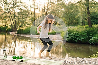 Delightful little female athlete happily jumping on green mat on sandy beach near river. Girl doing sports outdoors. Stock Photo