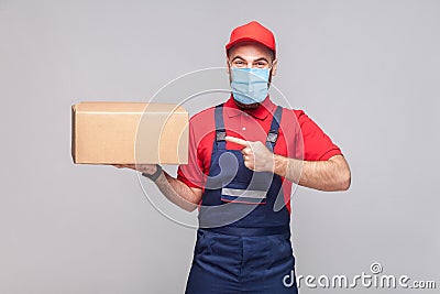 Delivery on quarantine. Young man with surgical medical mask in blue uniform and red t-shirt standing, holding and pointing finger Stock Photo