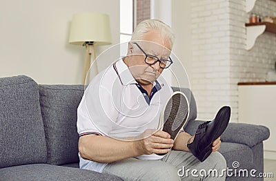 Demented retired senior man sitting on couch and choosing which type of shoes to wear Stock Photo