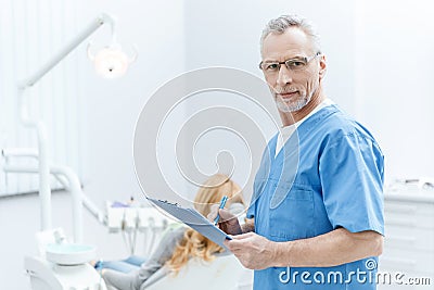 Dentist in uniform writing on clipboard in dental clinic with patient behind Stock Photo