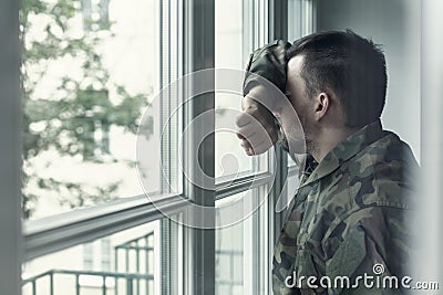 Depressed and sad soldier in green uniform with trauma after war standing near the window Stock Photo