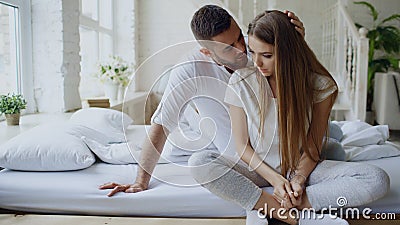 Depressed young woman sitting in bed and crying while her boylfriend come and embrace her and kiss in bedroom at home Stock Photo