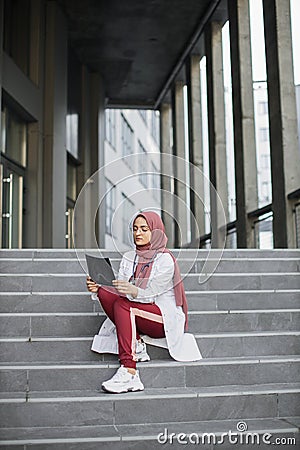 Muslim doctor looking at xray scan of patient's skull, sitting on the stairs of modern hospital Stock Photo