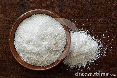 Desiccated coconut in dark wooden bowl isolated on dark brown wood from above. Spilled coconut. Stock Photo