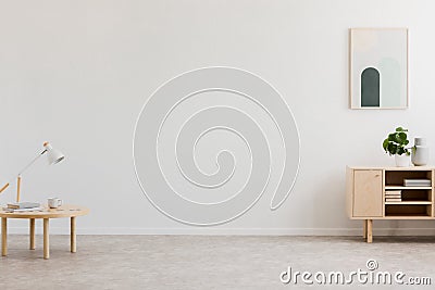 Desk lamp on a small table and a simple, wooden cabinet in an empty living room interior with white wall and place for a sofa. Rea Stock Photo