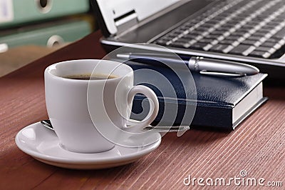 Desktop with coffee cup, opened laptop computer, diary, pan and document folders on background, no people, focused on coffee Stock Photo