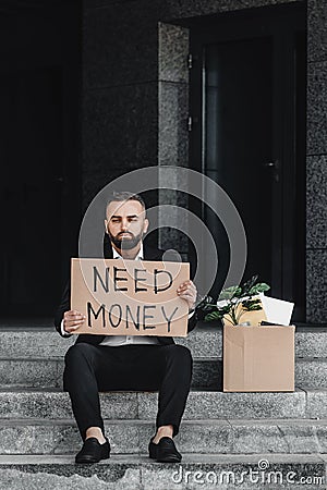 Despaired middle-aged businessman holding cardboard sign with need money text, sitting near business centre, copy space Stock Photo