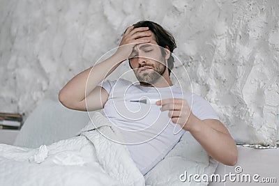 Despaired millennial caucasian guy with stubble on bed, touches forehead, looks at thermometer in bedroom Stock Photo