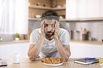 Despaired tired millennial caucasian man with beard holds head with hand, asleep at table with croissant Stock Photo