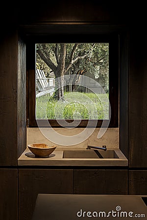 Detail of contemporary dark wooden kitchen with a sink beside a bowl. The garden-facing window offers a view of olive trees Stock Photo