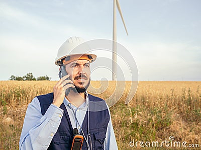 Detail of a male wind engineer with a safety helmet talking on the phone in a field of windmills. Renewable energy Stock Photo