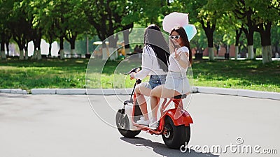 Deux belles filles dans des shorts de denim montent en parc sur un scooter électrique pendant l'été banque de vidéos Deux belles filles dans des shorts de denim montent en parc sur un scooter électrique pendant l'été banque de vidéos