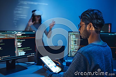 developer uses a VR headset to interact with projected interface designs, while colleagues discuss and code on screens, Stock Photo